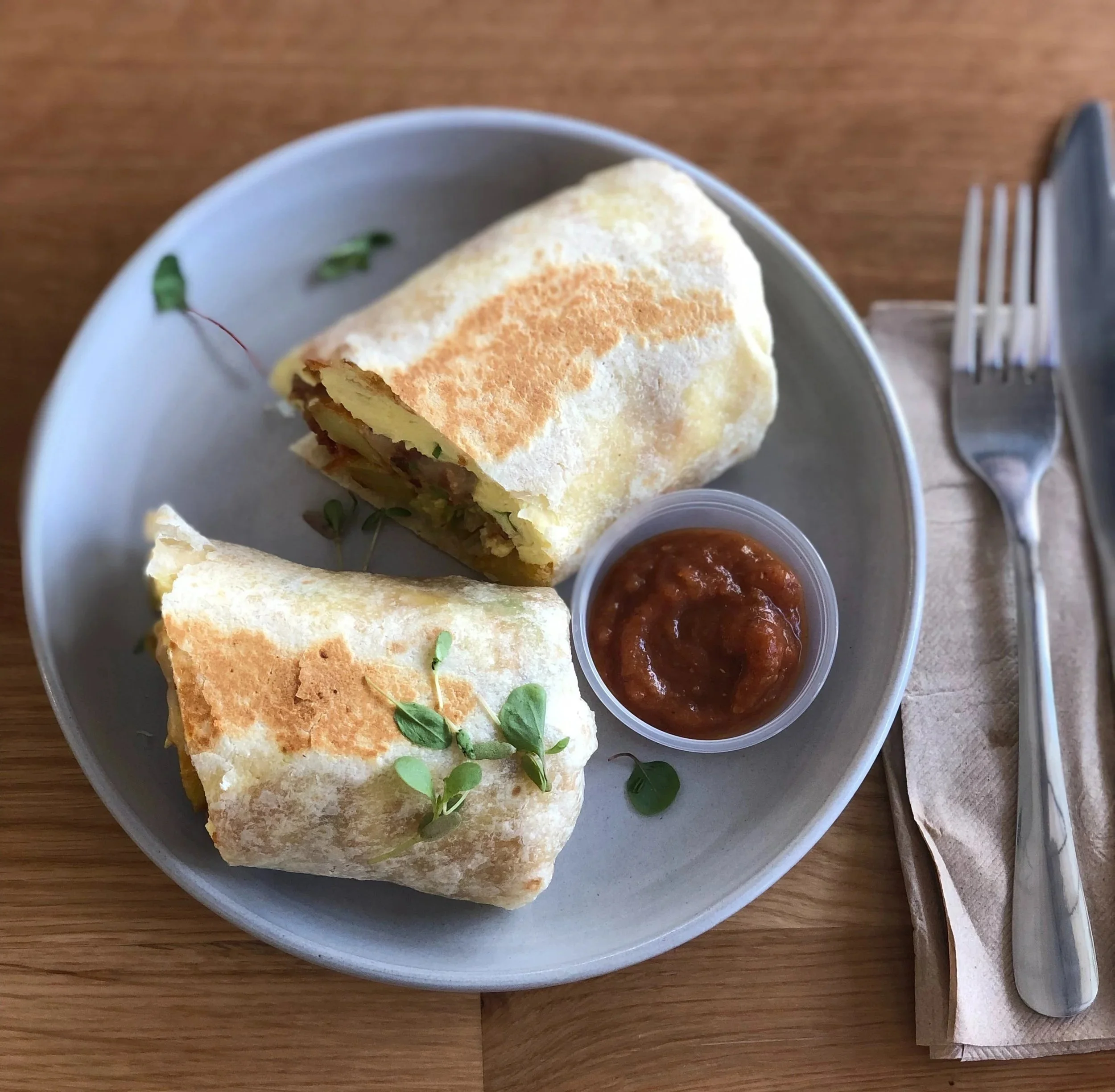 A white plate with a wrapped sandwich, side of salsa, and foil-wrapped utensil on a wooden table.