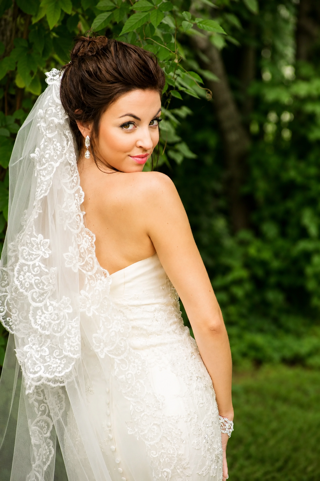 A bride in a strapless white wedding dress with a lace veil inside a green outdoor setting.