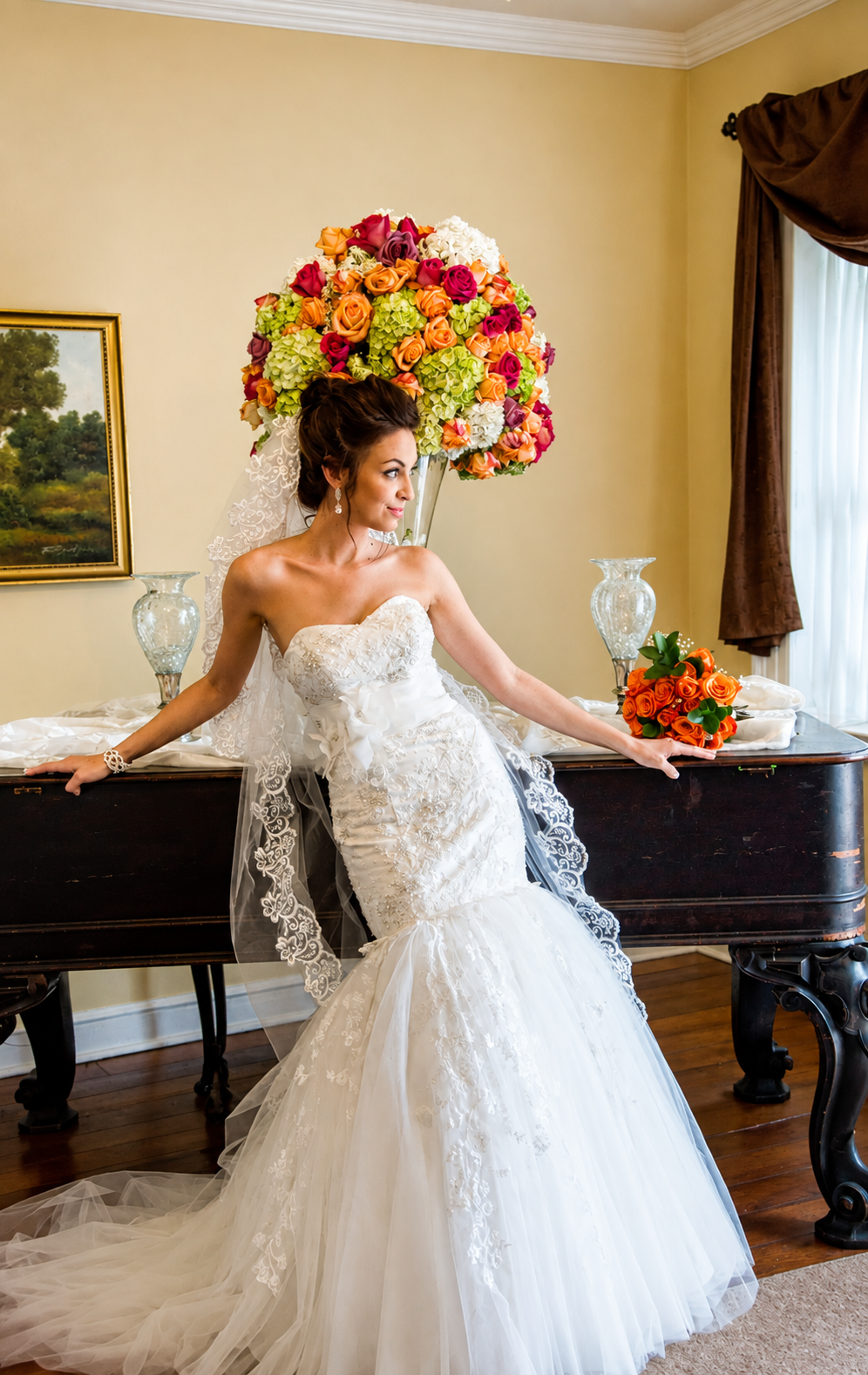 A bride in a strapless, lace wedding gown with intricate embroidery, standing in a room with a wooden sideboard. Behind her, a large floral arrangement with multicolored roses and hydrangeas. She is holding a bouquet of orange roses and looking to th