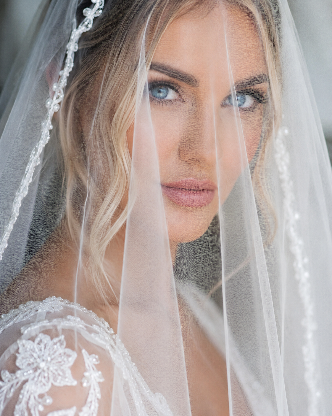 Close-up of a bride with curly blonde hair, wearing a lace wedding dress and a sheer veil, looking directly at the camera.