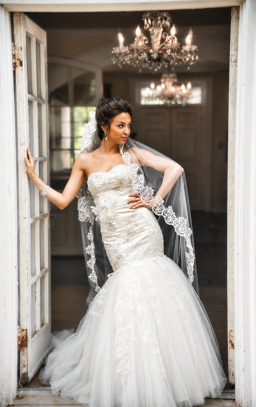 Bride in a strapless wedding gown with lace details and a tulle skirt, standing in a doorway with chandeliers visible inside in the background.