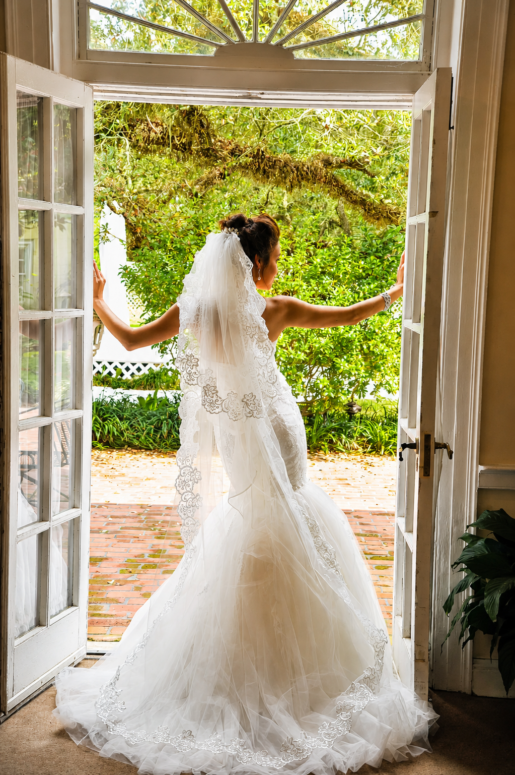 A bride in a white wedding gown and veil stands at open French doors, gazing outside at green foliage and brick patio, with arms outstretched holding the doorframes.