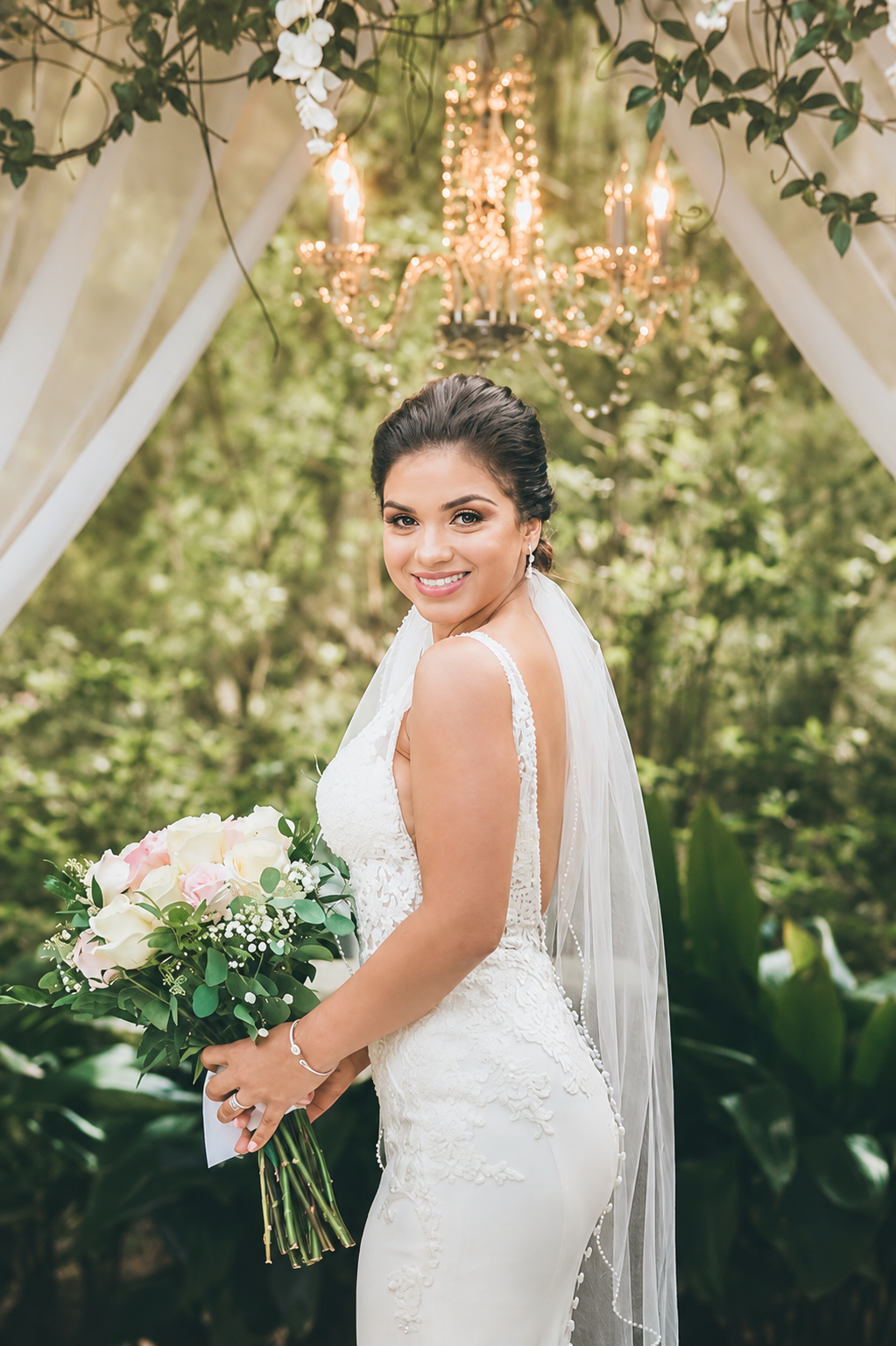 A bride with dark hair in a white lace wedding gown holding a bouquet of pink and white roses, standing outdoors under a decorated arch with a chandelier and greenery.