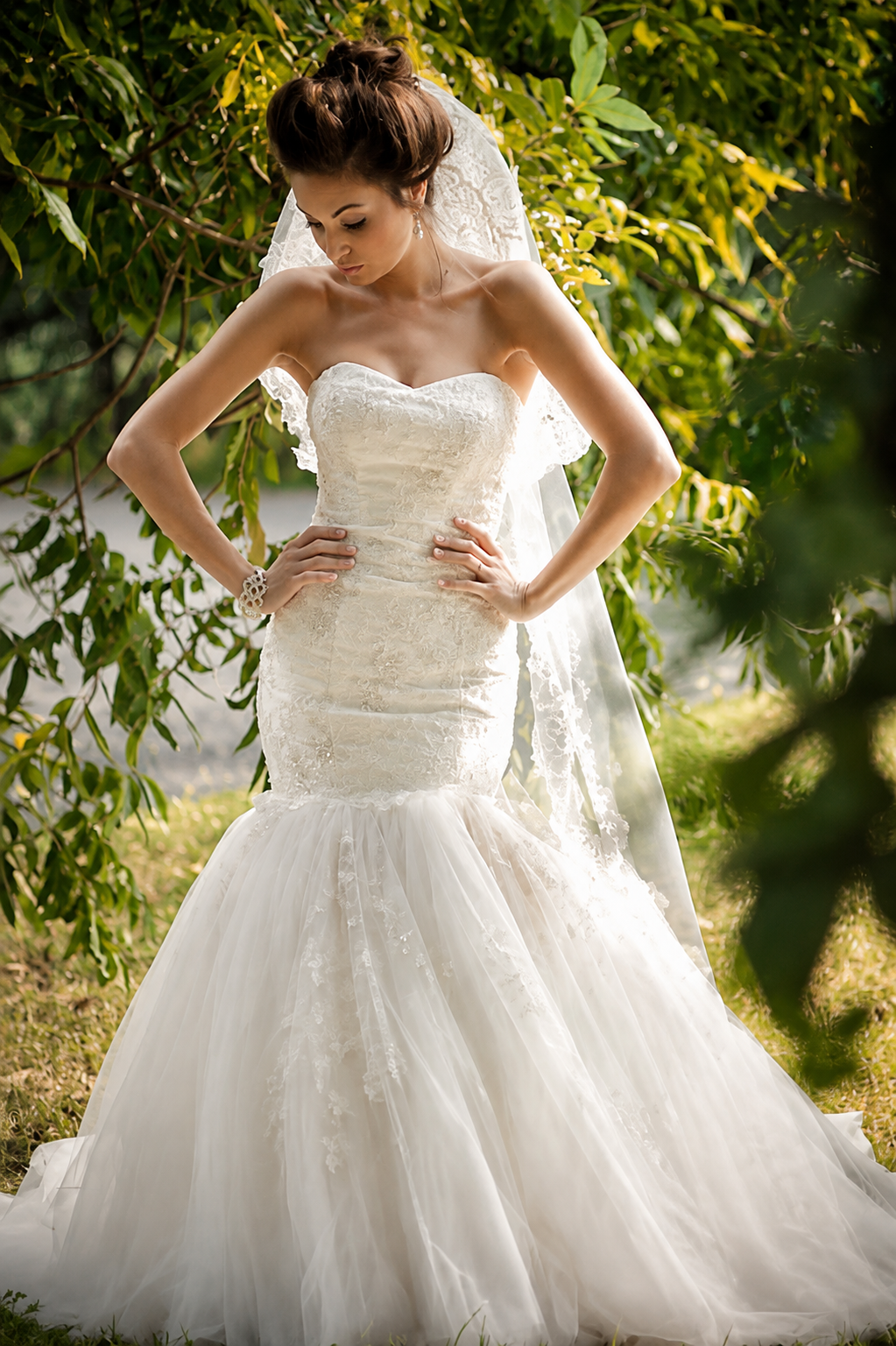 A bride in a strapless lace wedding gown with a full tulle skirt, standing outdoors among green foliage, with hands on her hips and looking down.