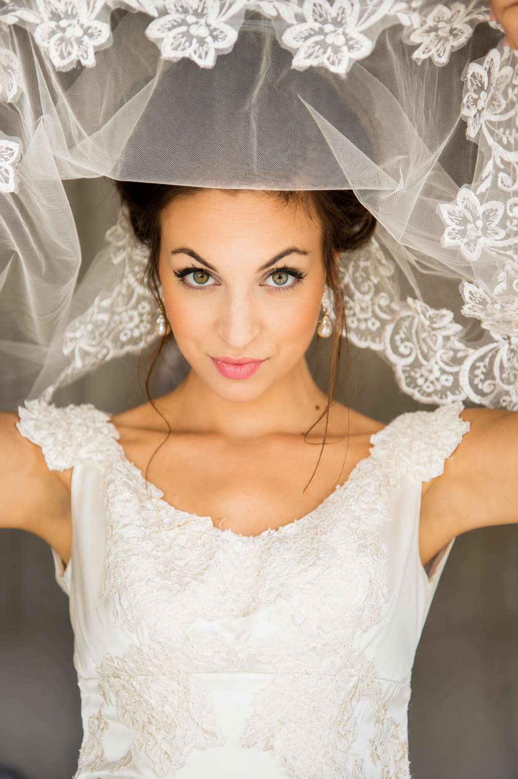 A bride with green eyes and dark hair, wearing pearl earrings and a white lace wedding dress, lying under a lace veil with floral embroidery.