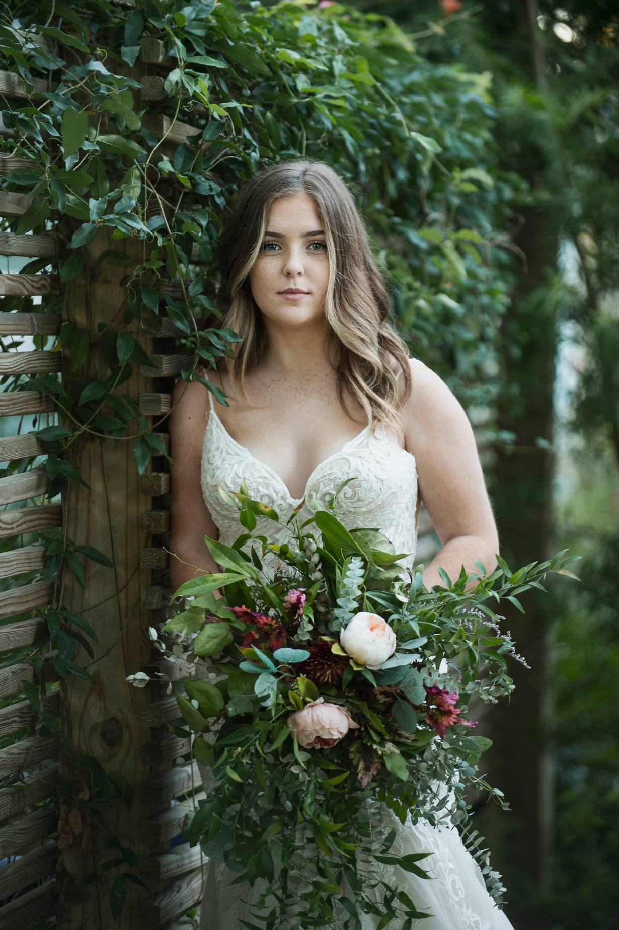 A woman in a white wedding dress holding a bouquet of flowers, standing outdoors next to a wooden fence covered in green foliage.