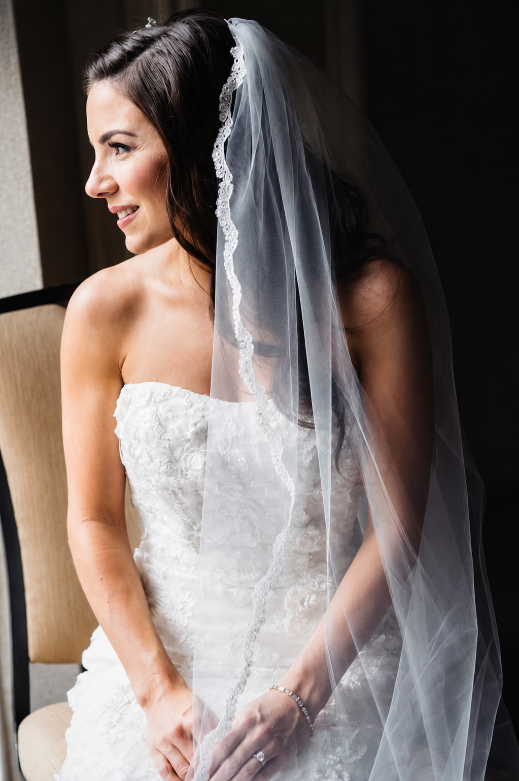A bride with dark brown hair and a white wedding dress sitting on a chair, holding her right hand with a diamond ring, and smiling while looking to the side. She is wearing a veil with lace edges and a bracelet.