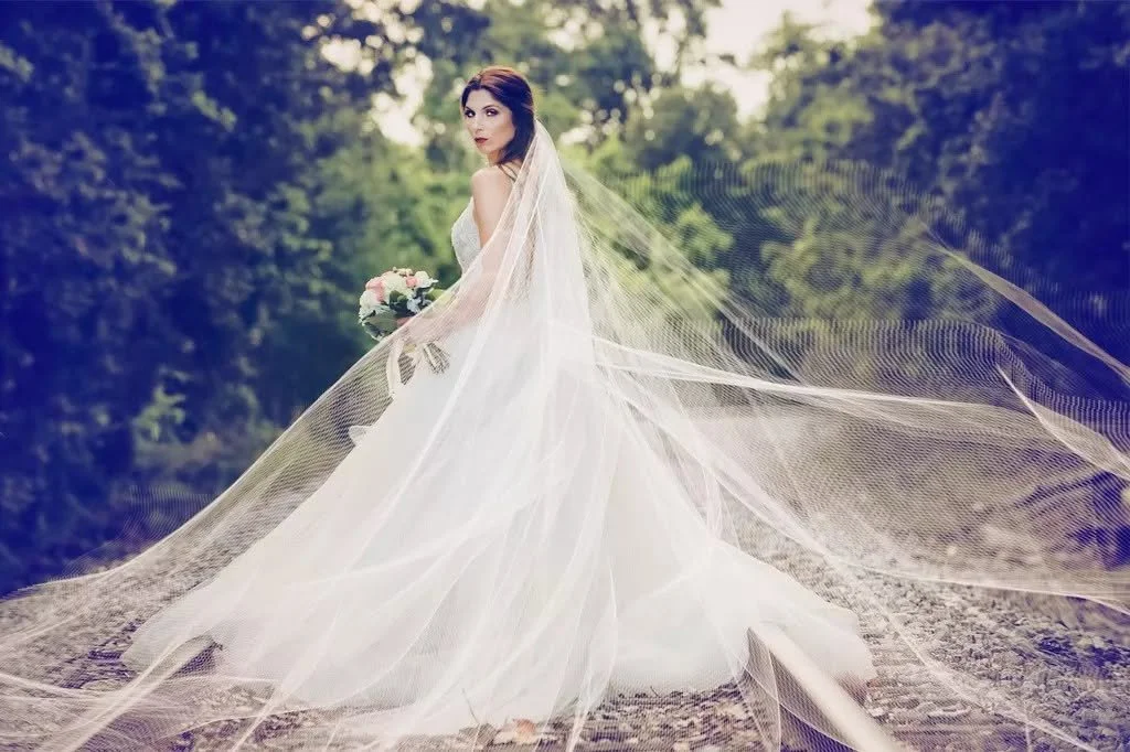 Bride in a white wedding gown with a long veil holding a bouquet of flowers in an outdoor setting with trees in the background.
