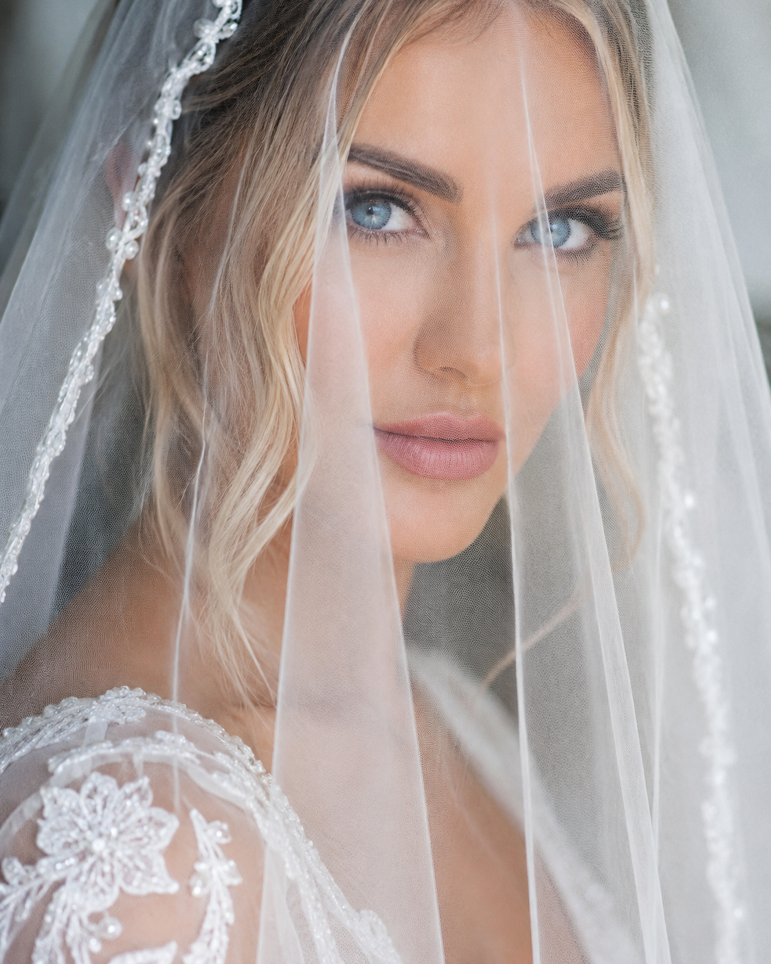 A bride with blonde hair and blue eyes wearing a wedding dress with lace details, covered partially by a sheer veil with embroidered embellishments.