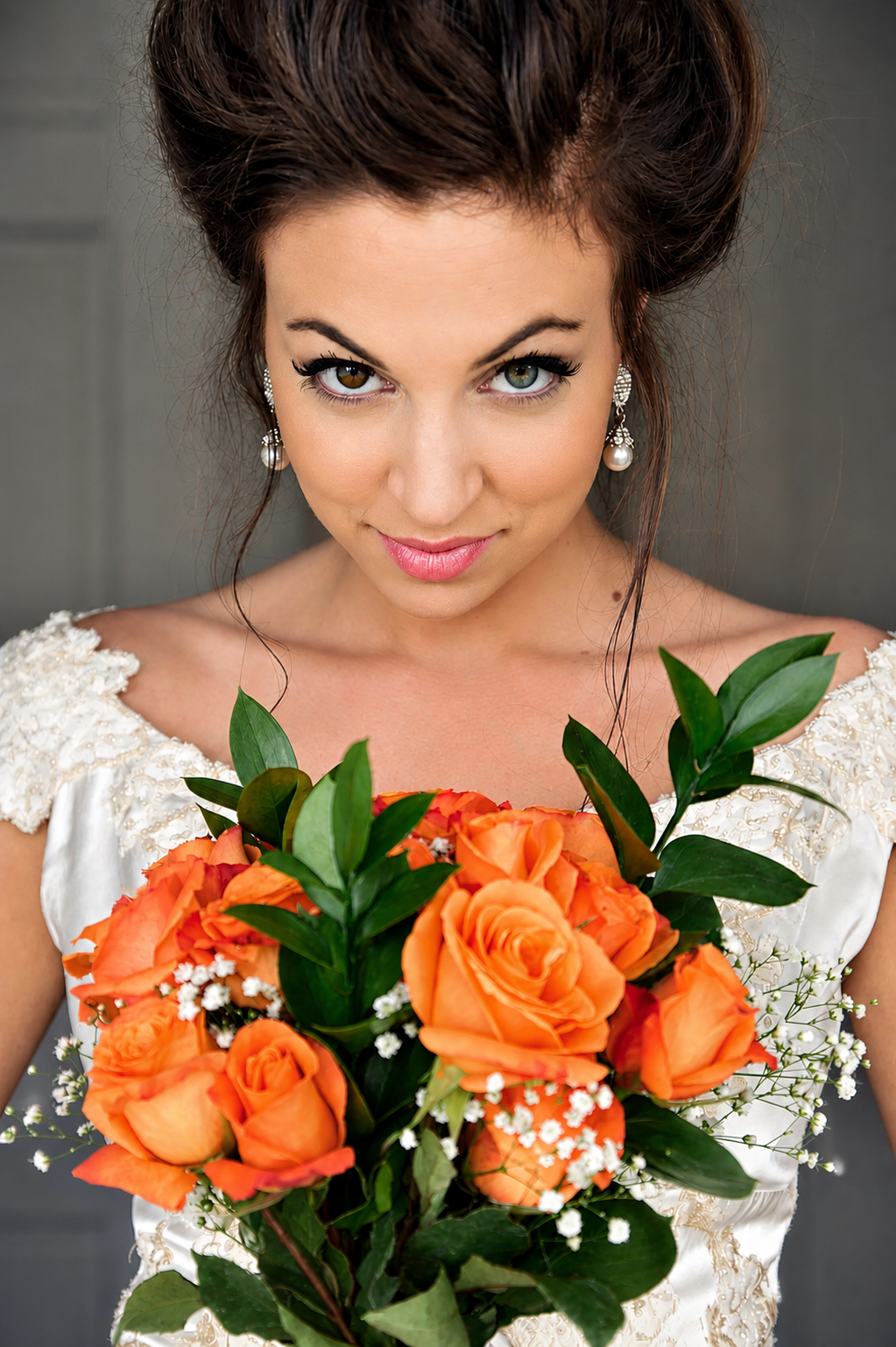 A woman in a white wedding dress holding a bouquet of orange roses, looking directly at the camera.