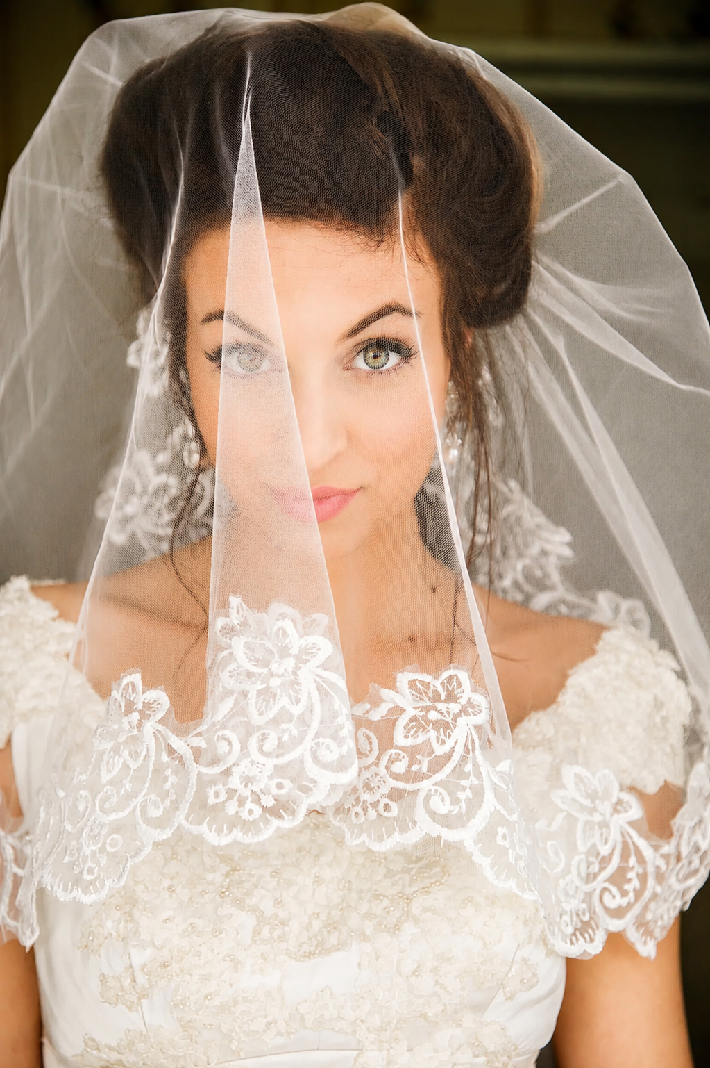 A bride with green eyes and dark hair partially covered by a lace veil, wearing a white wedding dress with floral lace details.