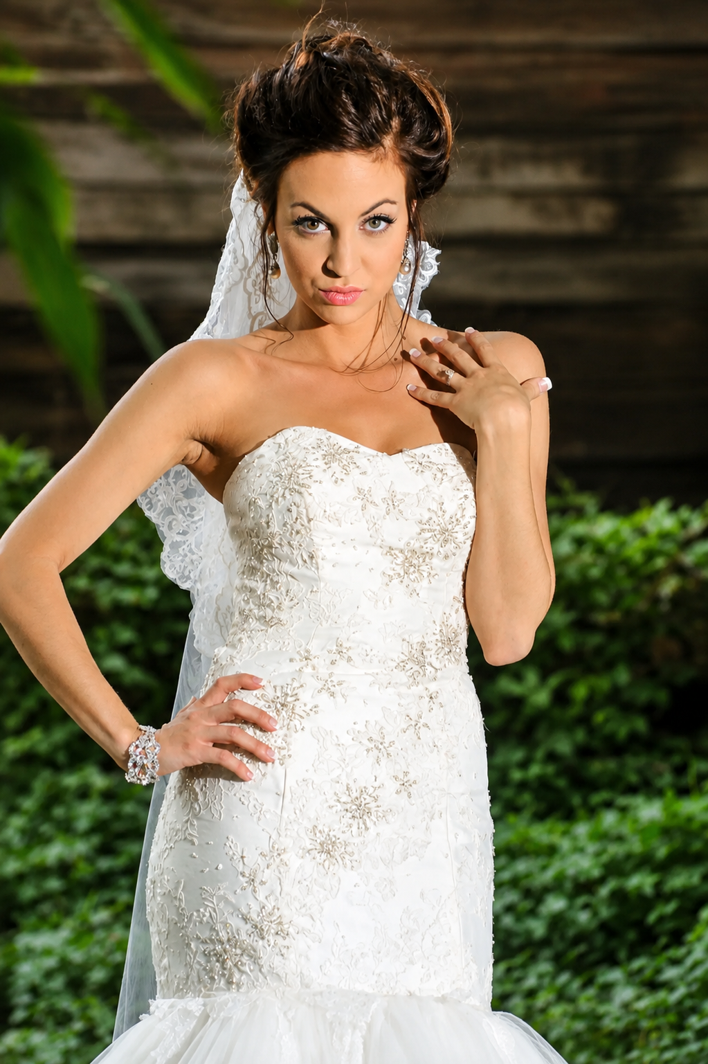 A woman in a white wedding dress with embroidery, wearing a bracelet, and a lace veil, posing outdoors with greenery and wooden background.