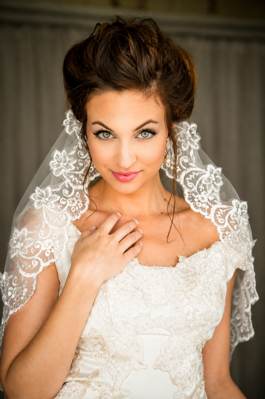 A bride in a white wedding dress with intricate lace details, wearing a lace veil with floral embroidery, touching her shoulder, looking at the camera with a confident expression.