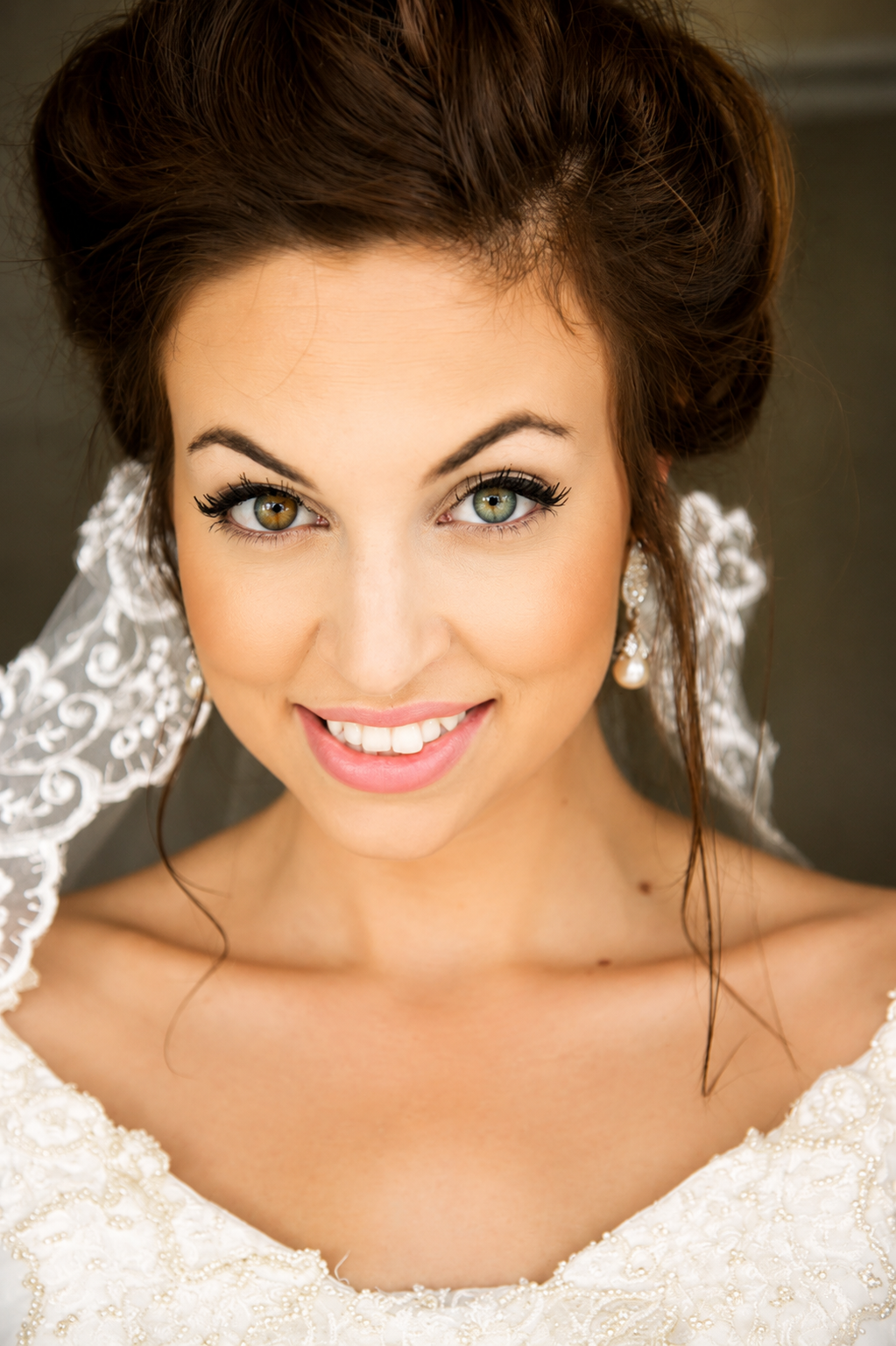 Close-up of a smiling bride with green eyes and dark brown hair styled in an updo, wearing pearl earrings and a white lace wedding dress.
