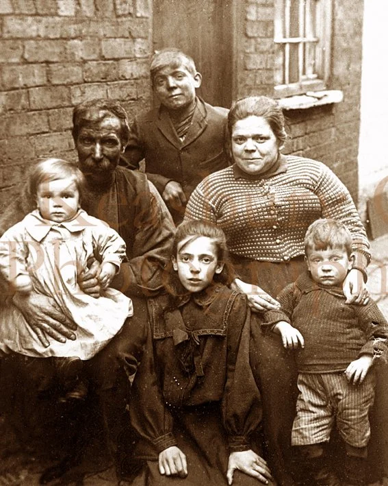 A vintage black-and-white photo of six people, including three adults and three children, gathered outdoors against a brick wall, with a window visible behind them. The scene appears to be from an earlier time period.