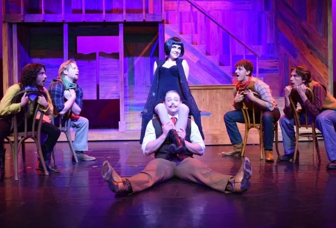 Theater scene with six actors on stage, one woman standing above a man sitting with legs spread, surrounded by four men seated on chairs, all in theatrical costumes, with a wooden set background and stage lighting.