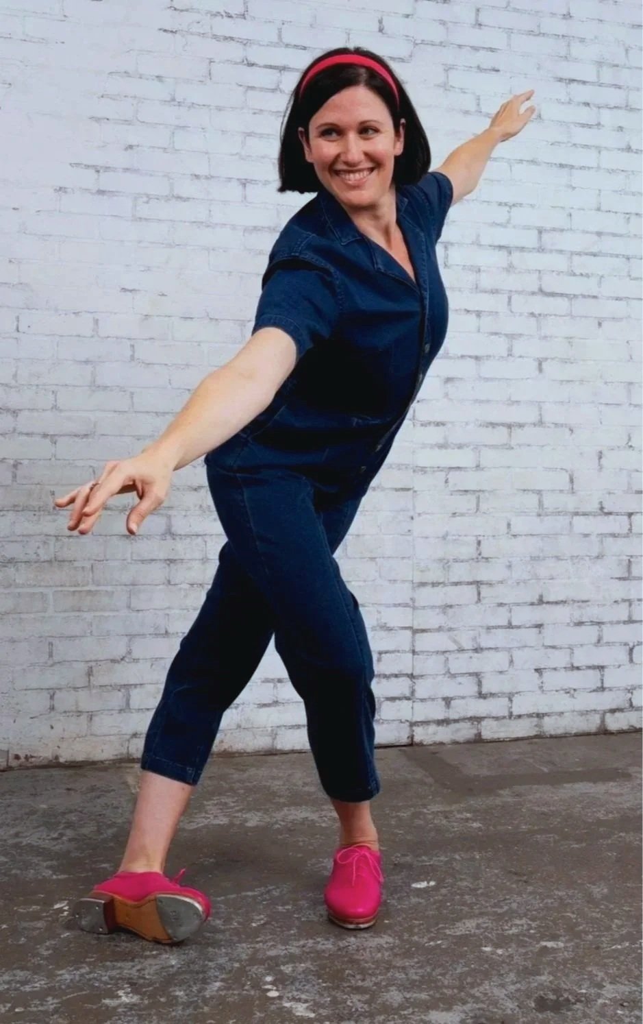 Woman with black hair, pink headband, blue denim shirt, and pink shoes striking a playful dance pose in front of a white brick wall.