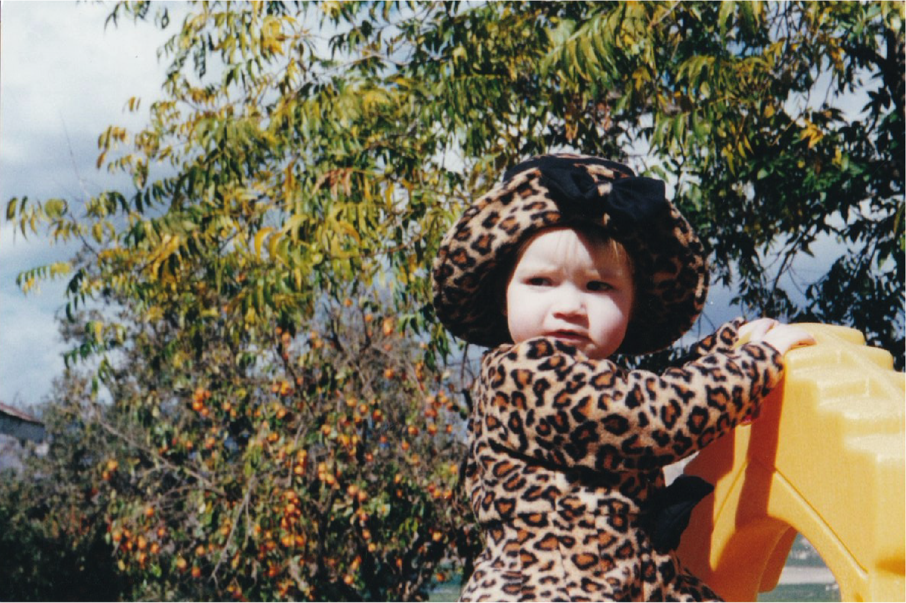 Young child in leopard print outfit and hat playing outdoors, holding a yellow object, with trees and sky in the background, on the ranch.