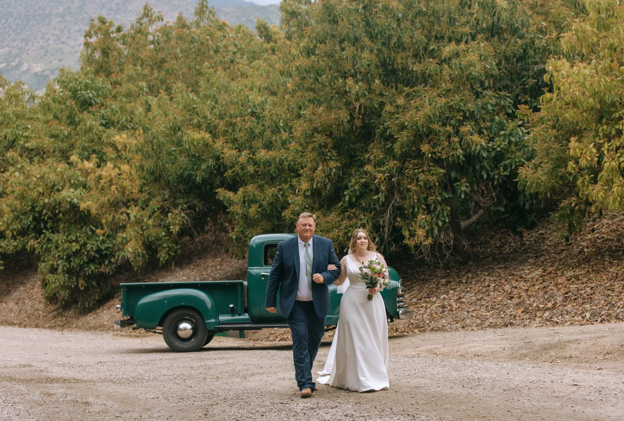 A bride and her father walking together in front of a green pickup truck, surrounded by large trees.