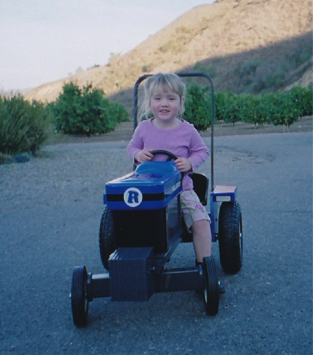 A young girl with blonde hair, wearing a pink long-sleeve shirt and patterned pants, sitting in tractor on an outdoor area with green bushes and on the ranch.