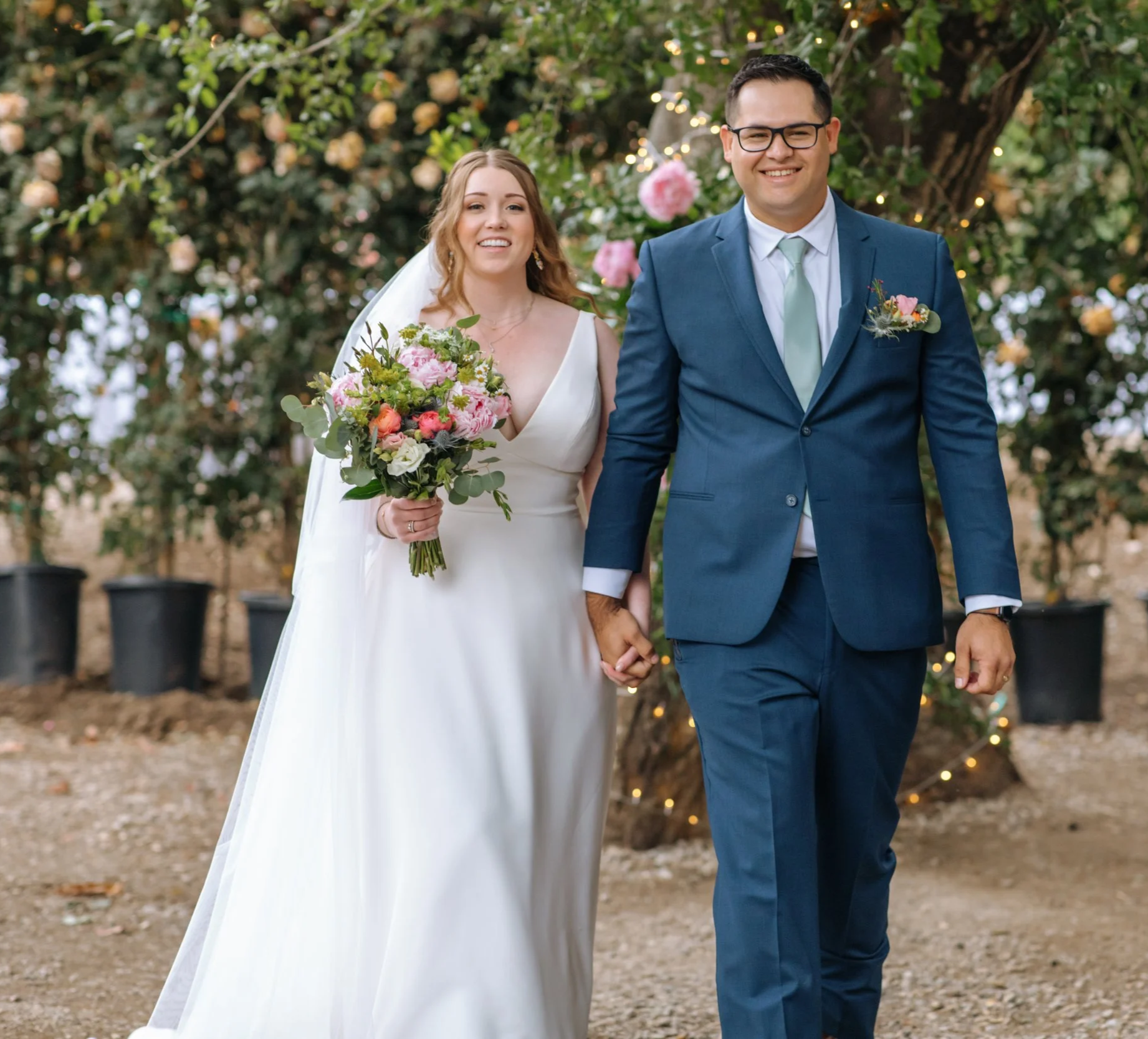 A bride and groom holding hands at their outdoor wedding, surrounded by greenery and flowers, with soft lighting on the ranch.
