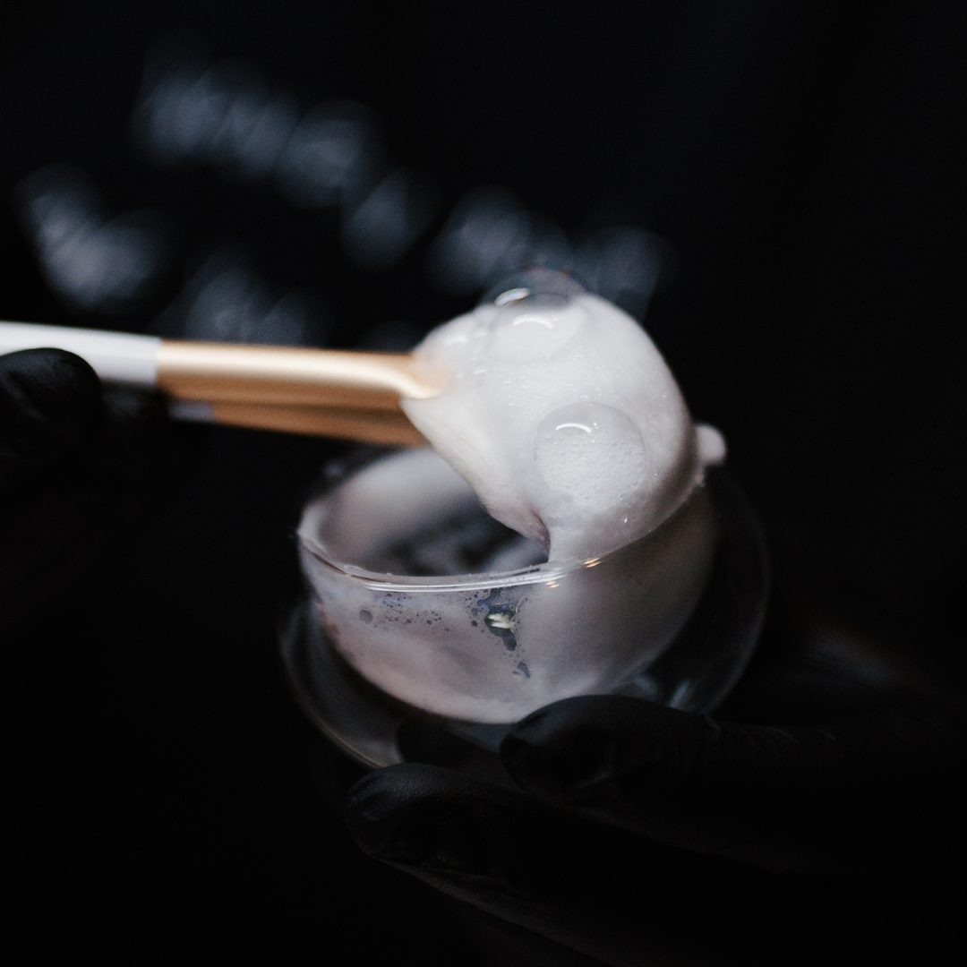 Close-up of a person wearing black gloves holding a small glass container, with a soft fan brush  applying a white, foamy facial cleanser