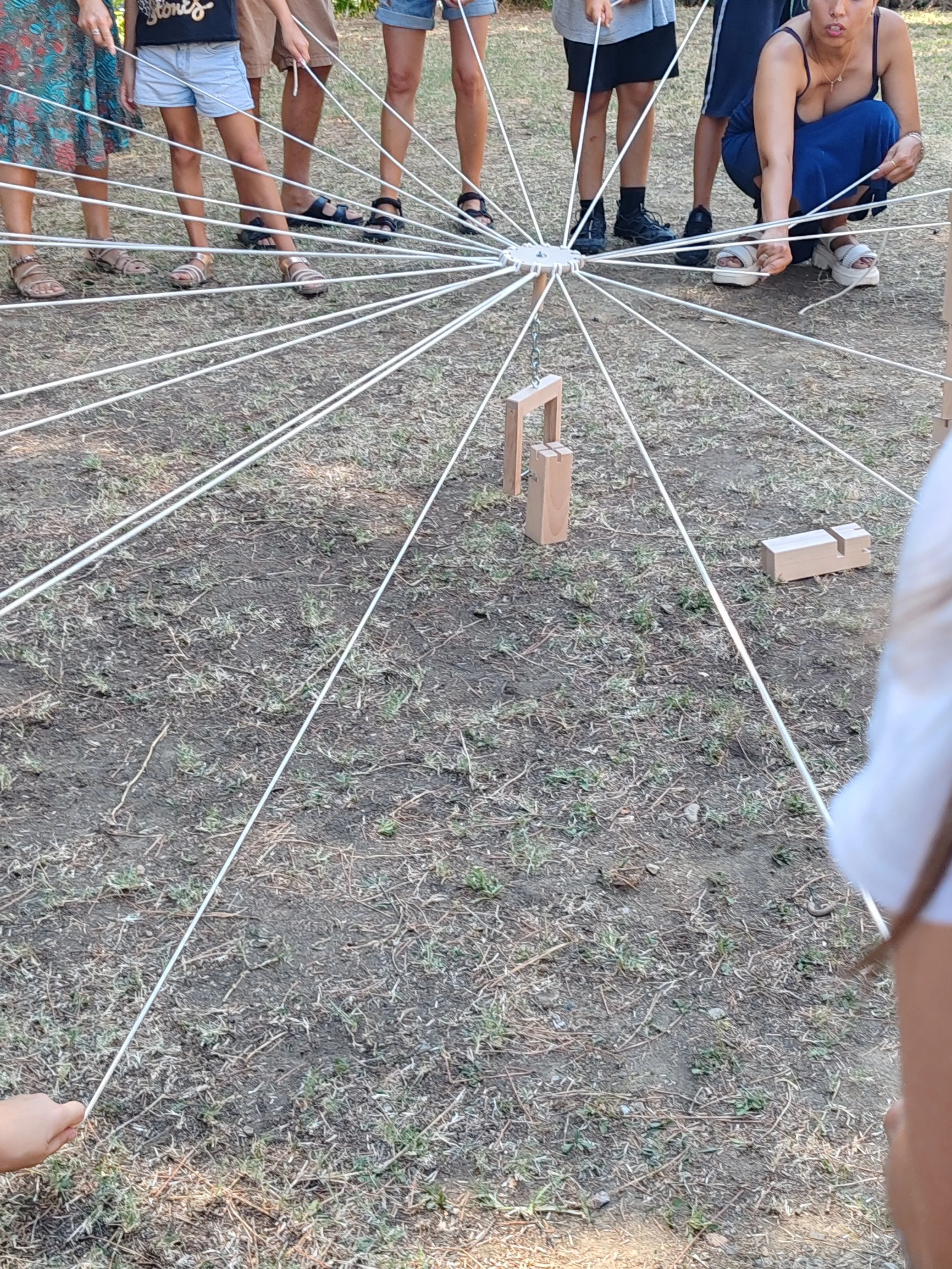 Group of people standing and squatting around a spider web-like structure made of strings with wooden blocks on the ground, outdoors on dirt ground.