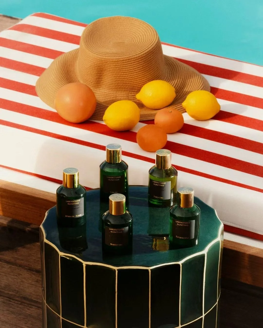 A straw hat, lemons, and onions on a striped red and white tablecloth, with bottles of perfume or essential oils on a dark green table with gold accents.