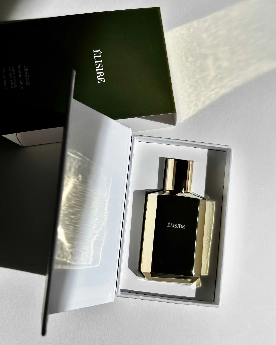 A black and gold perfume bottle labeled 'ÉLISIRE' inside a white box, with a green box labeled 'ÉLISIRE' beside it.