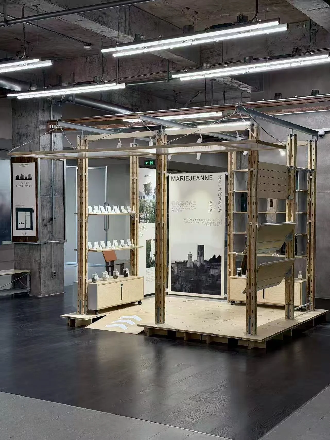 Exhibition booth with wooden frame and display shelves, featuring posters and small bottles, in an indoor setting with industrial ceiling and bright lighting.
