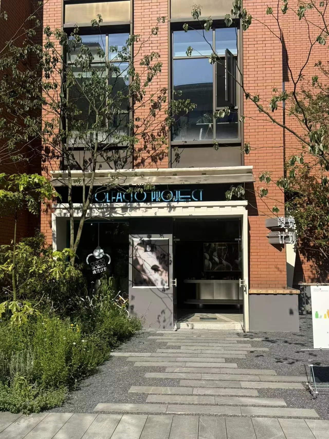 The entrance to a building with a sign that reads 'Olificio Project' in blue letters. The building has a red brick exterior, large windows, and a small garden with greenery in front.