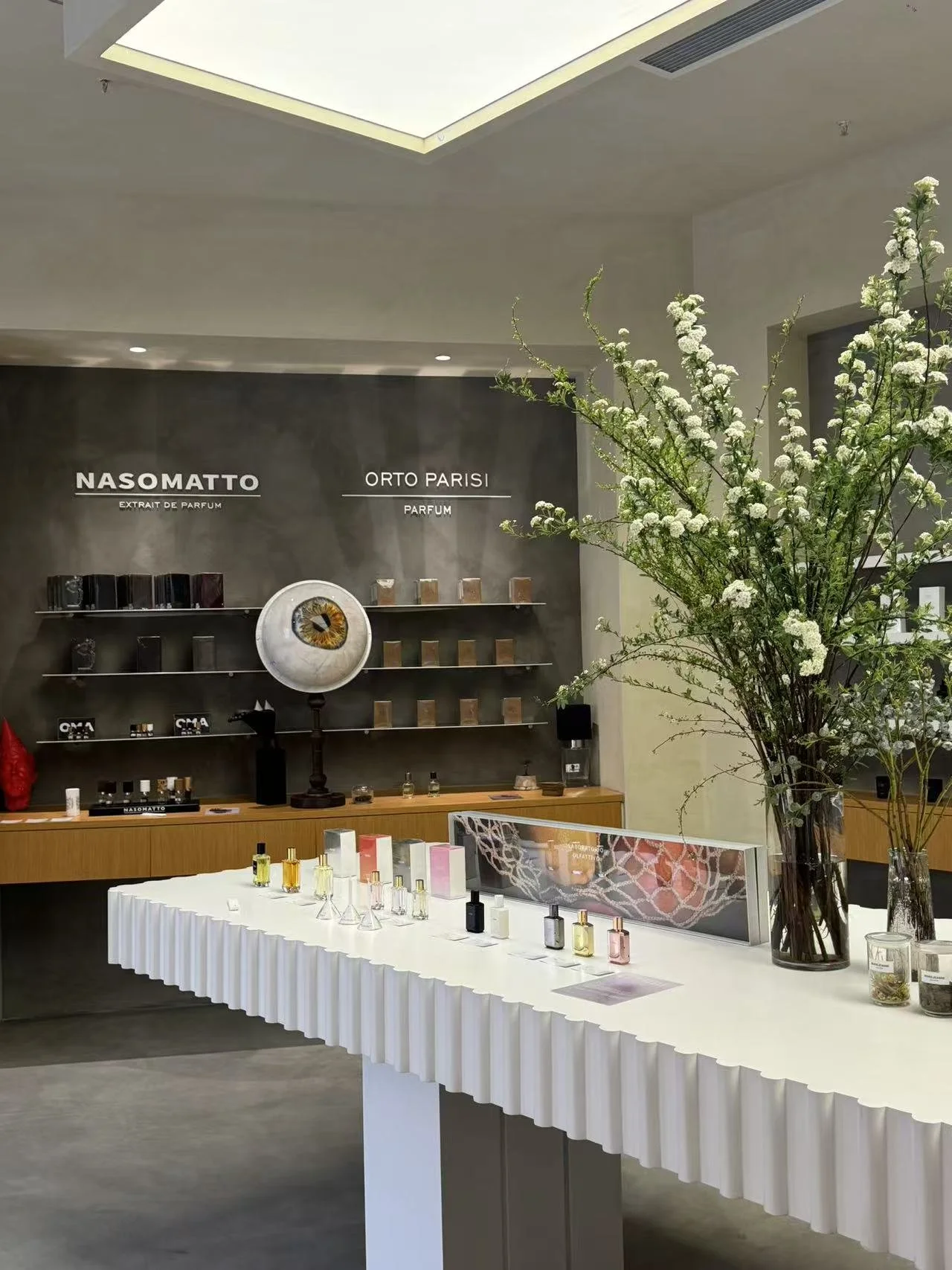 Display of perfume bottles on a white table with a slit-like pattern in a retail store, with large floral arrangement and wall shelving of perfumes behind.