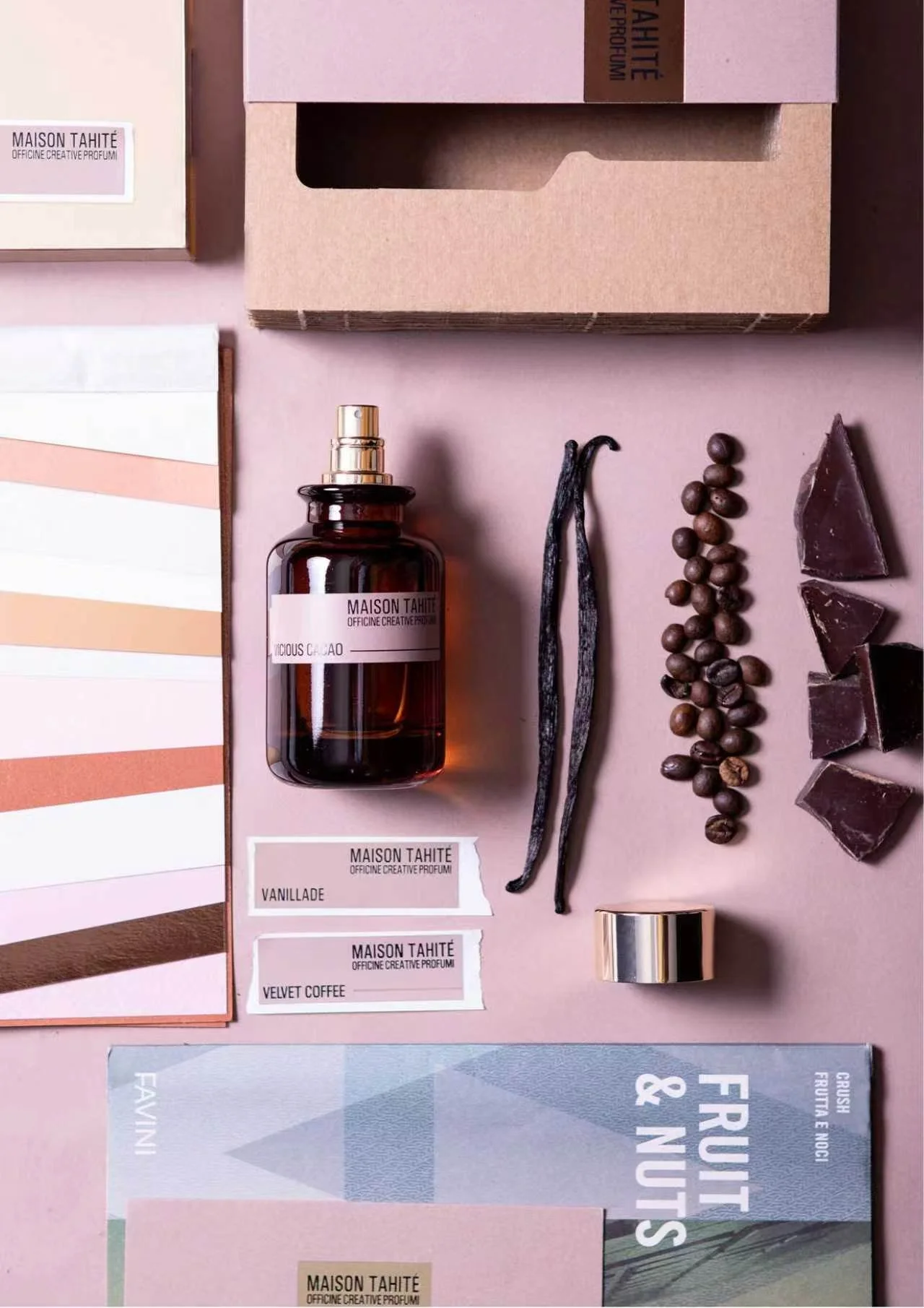 Flatlay of skincare and fragrance items on pastel pink background, including a brown glass spray bottle labeled 'MAISON TAHITÈ', vanilla pods, chocolate chunks, a color palette, and sample labels for scents 'Vanillée' and 'Velvet Coffee'.