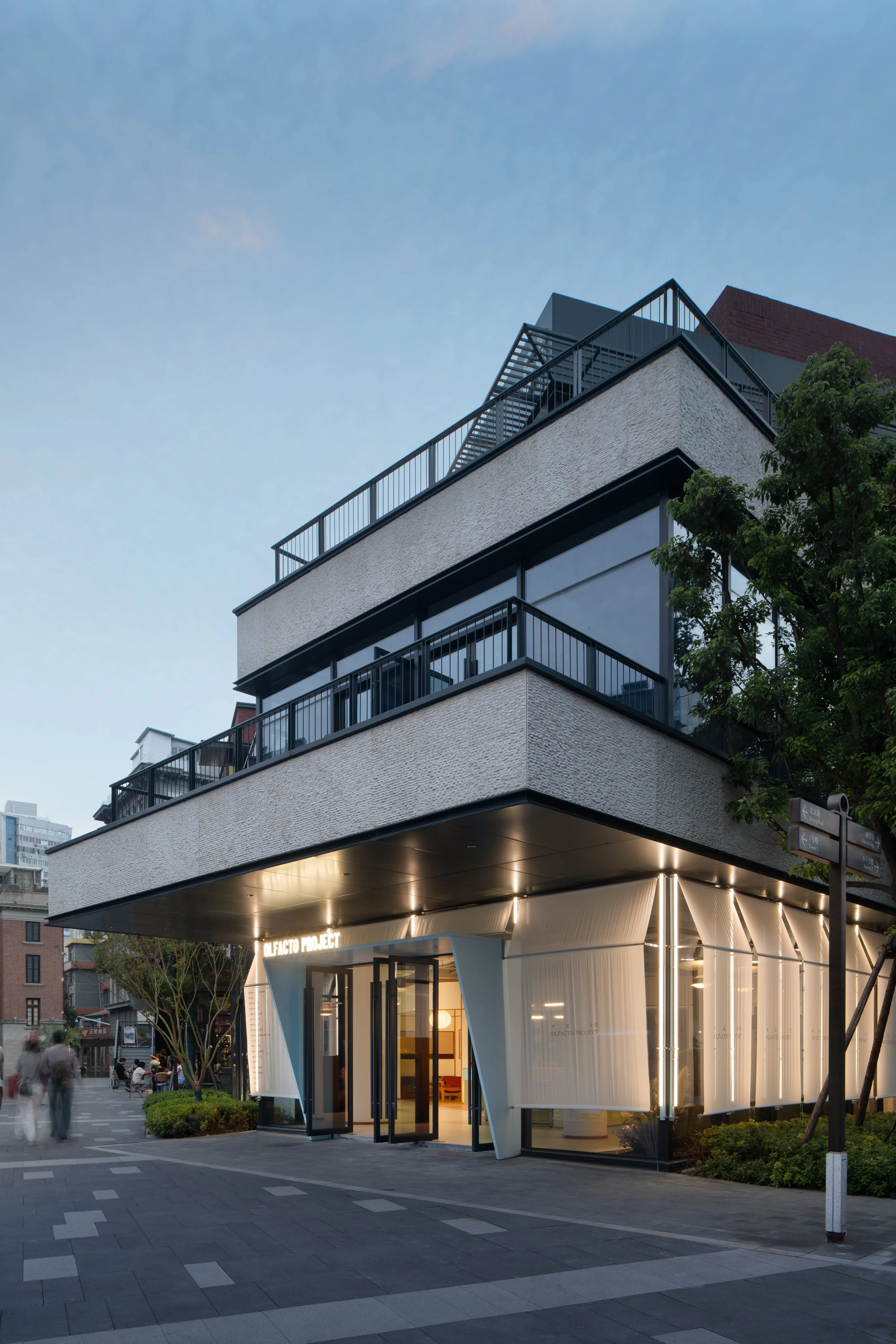 Modern building with a unique angular white façade and illuminated interior, located on a city street at dusk, with people walking nearby and trees lining the sidewalk.