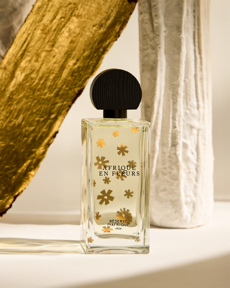 A perfume bottle labeled 'Afriqué En Fleurs' with gold flower designs, placed between a piece of wood and a textured ceramic vase.