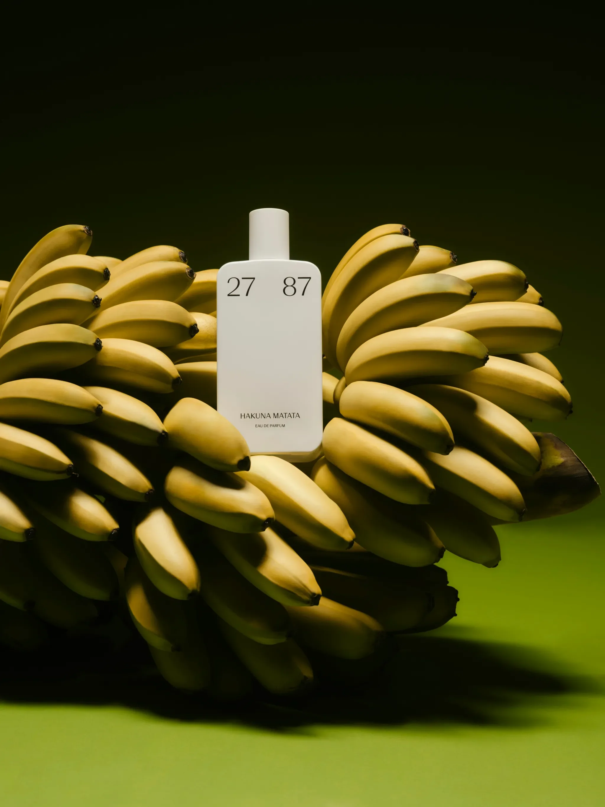 A white perfume bottle labeled 'Hakuna Matata' surrounded by ripe bananas on a green background.