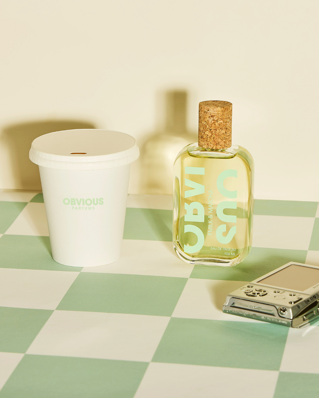 A bottle of OBVIOUS perfume, a white OBVIOUS Parfums cup, and a silver lighter on a green and white checkered surface with a light background.