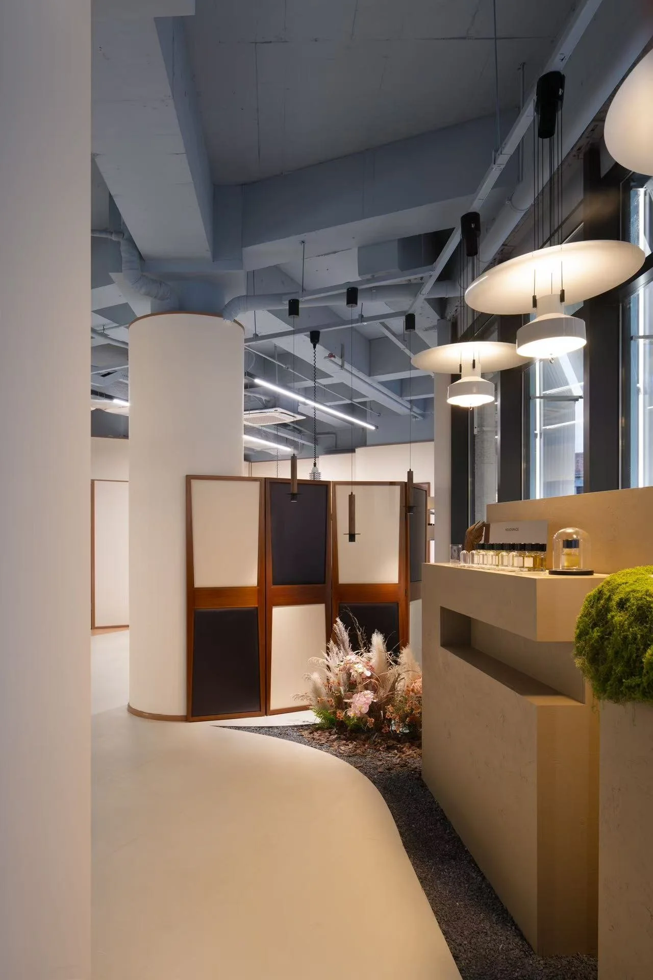 Interior of a modern retail or exhibition space with contemporary lighting, decorative room dividers, a curved counter, and floral arrangements.