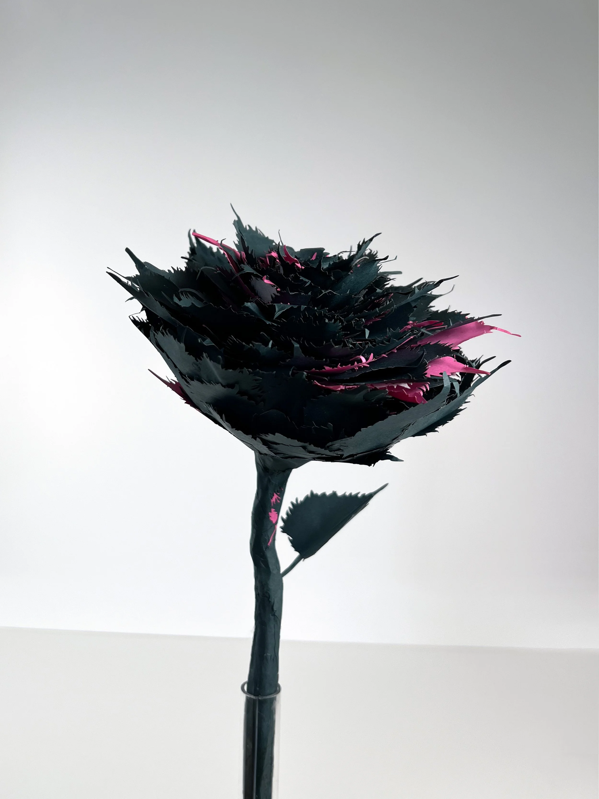 A black and pink artificial flower with jagged leaves and petals, mounted on a black stem, against a plain gray background.