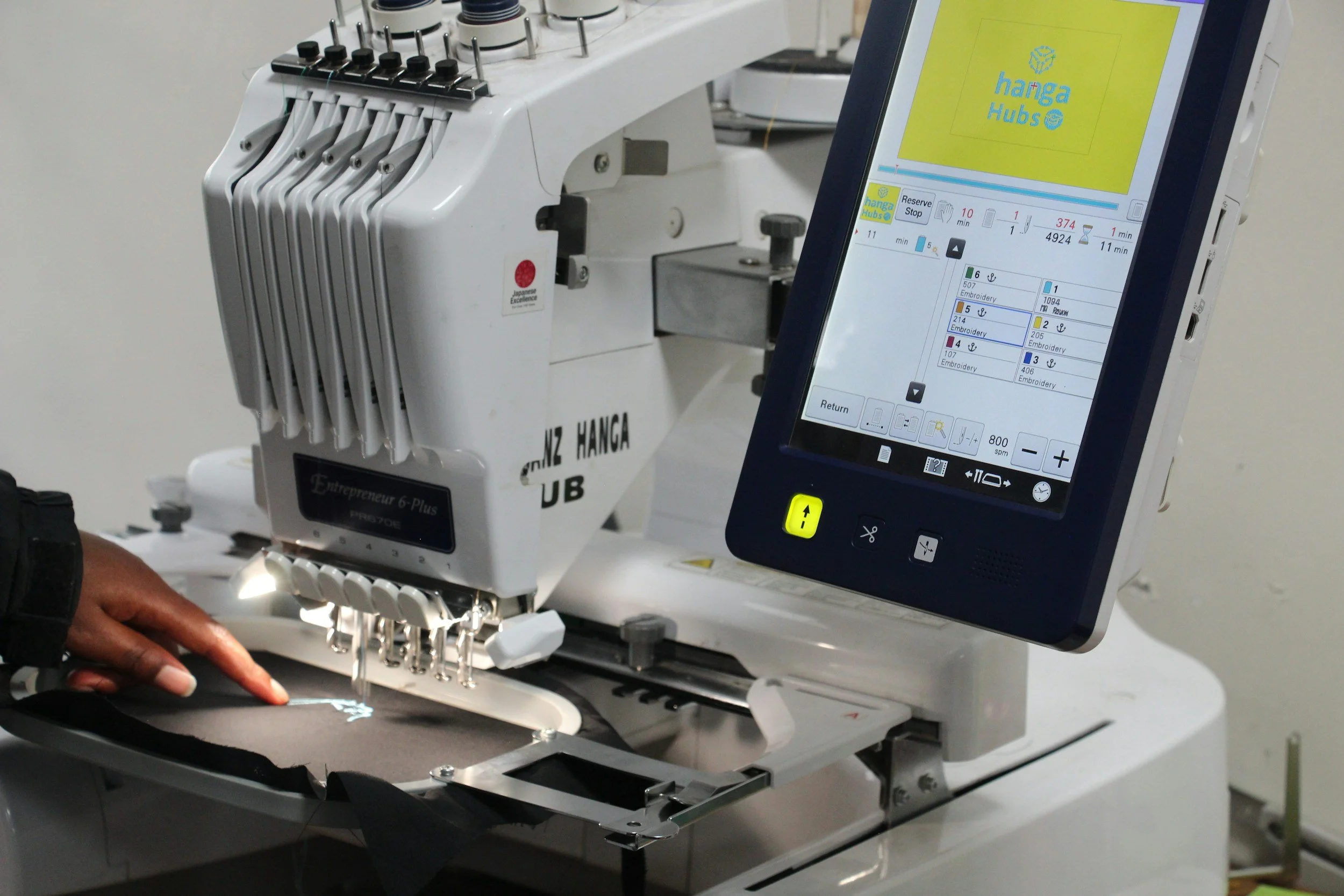 Close-up of a computer-controlled embroidery machine with a digital screen showing embroidery design options. The machine is actively stitching on a dark fabric.