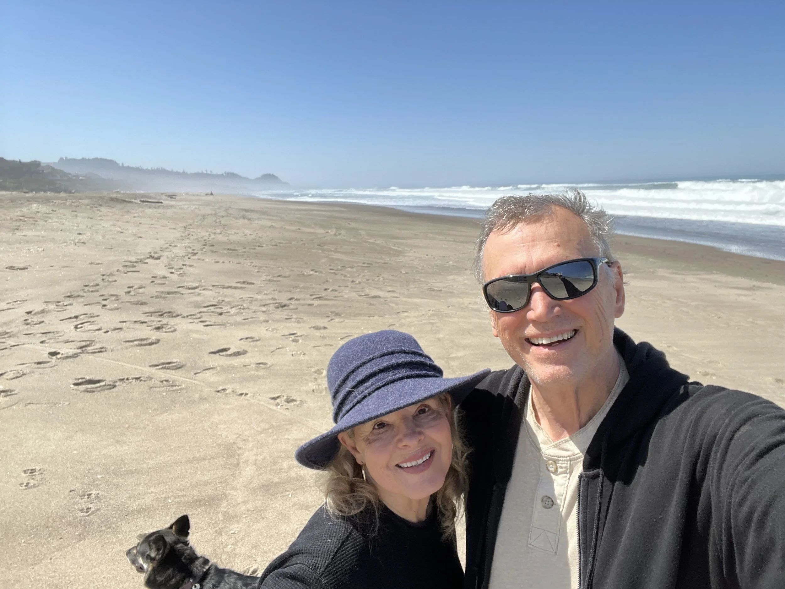 A man, a woman, and a small dog at the beach. The man is wearing sunglasses, the woman is wearing a sun hat, and all three are smiling. The beach has footprints in the sand, and the ocean waves are visible in the background under a clear blue sky.
