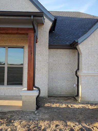 Partial view of a new residential house exterior showing a brick wall, window, wooden column, black gutters, and downspouts, with a dirt area in front.