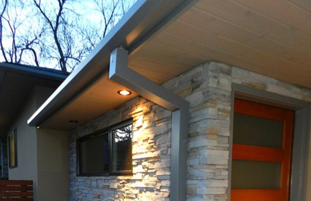 Close-up view of a house exterior showing a stone wall, window, door, and roof overhang with a ceiling light