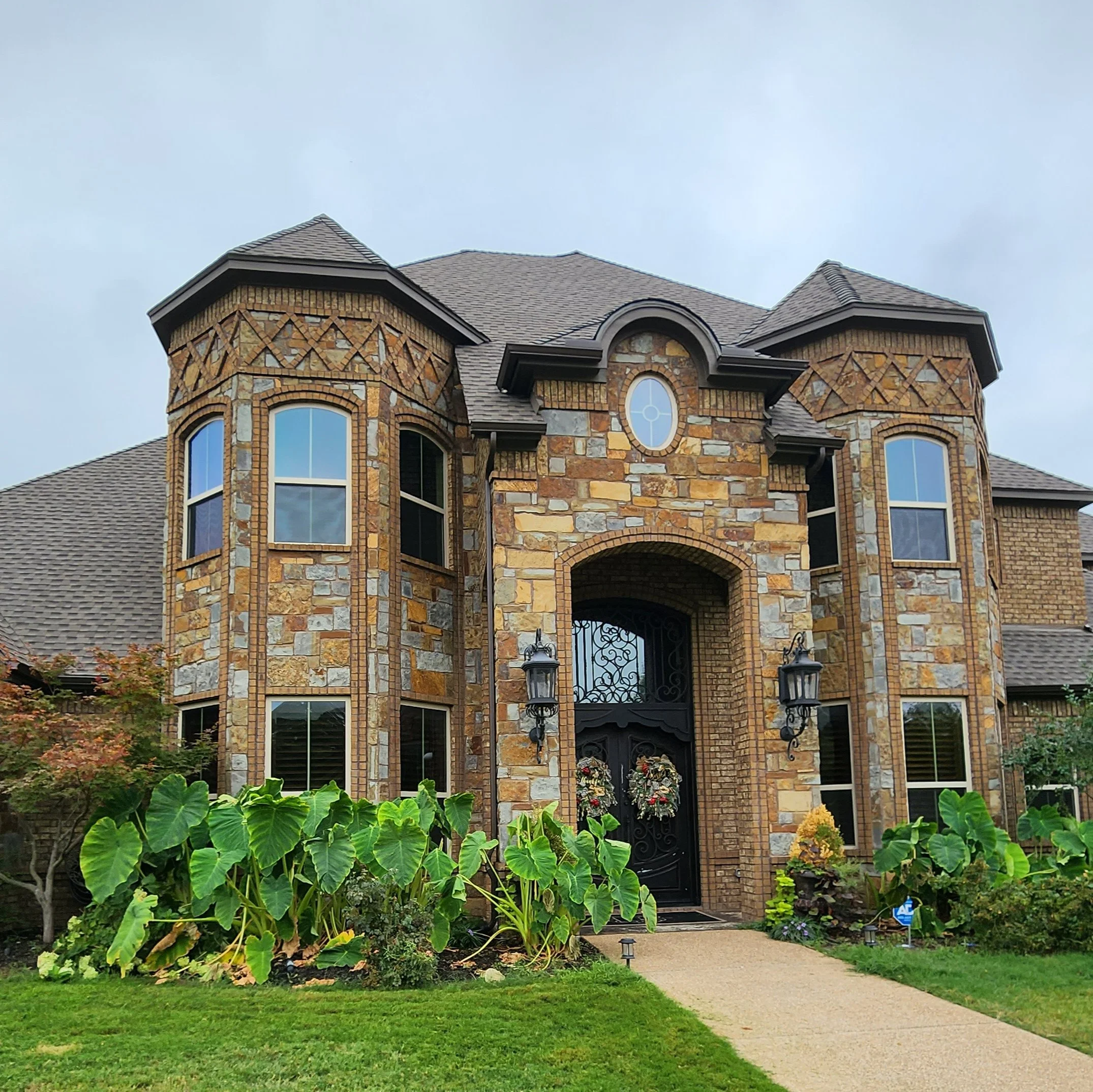 Front of a brick house with a stone facade, arched windows, and a black gated entrance, surrounded by greenery and plants.