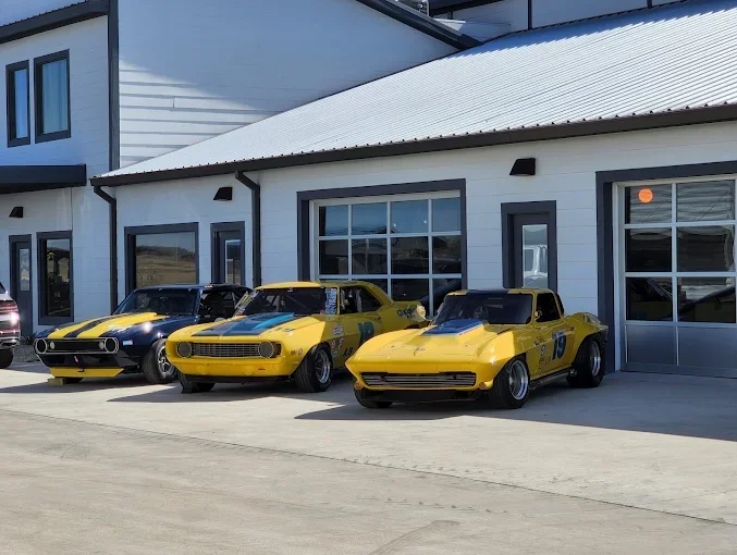 Three vintage race cars parked side by side in front of a white building with large windows. The cars are yellow with black accents.