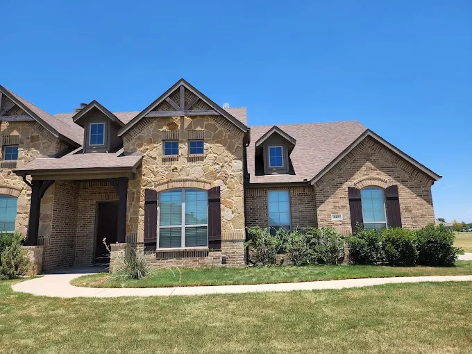 A large, two-story house with a brick and stone exterior, multiple gables, and a neatly landscaped front yard under a clear blue sky.