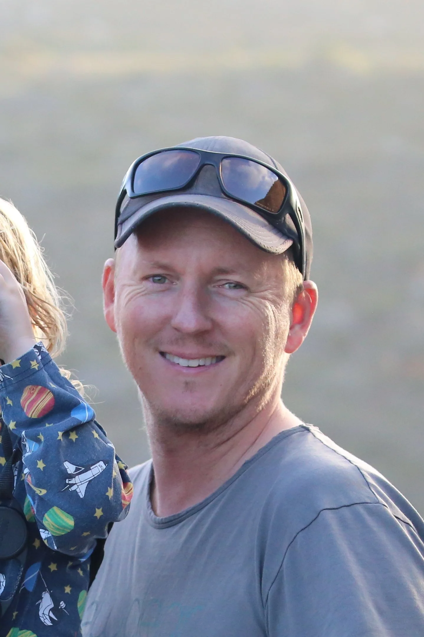 A smiling man with short hair wearing a gray t-shirt, sunglasses on his cap, holding a child wearing a blue jacket with colorful patterns, outdoors with a blurred cloudy sky in the background.
