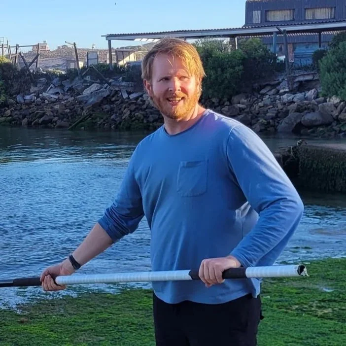 A man with red hair and beard outdoors near a body of water, holding a long pole, and smiling at the camera.
