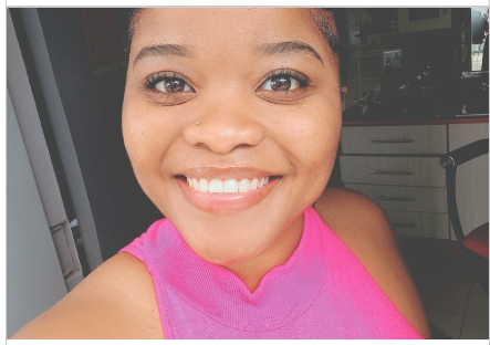 Close-up of a smiling woman with dark skin, wearing a pink sleeveless top, indoors with kitchen cabinets in the background.