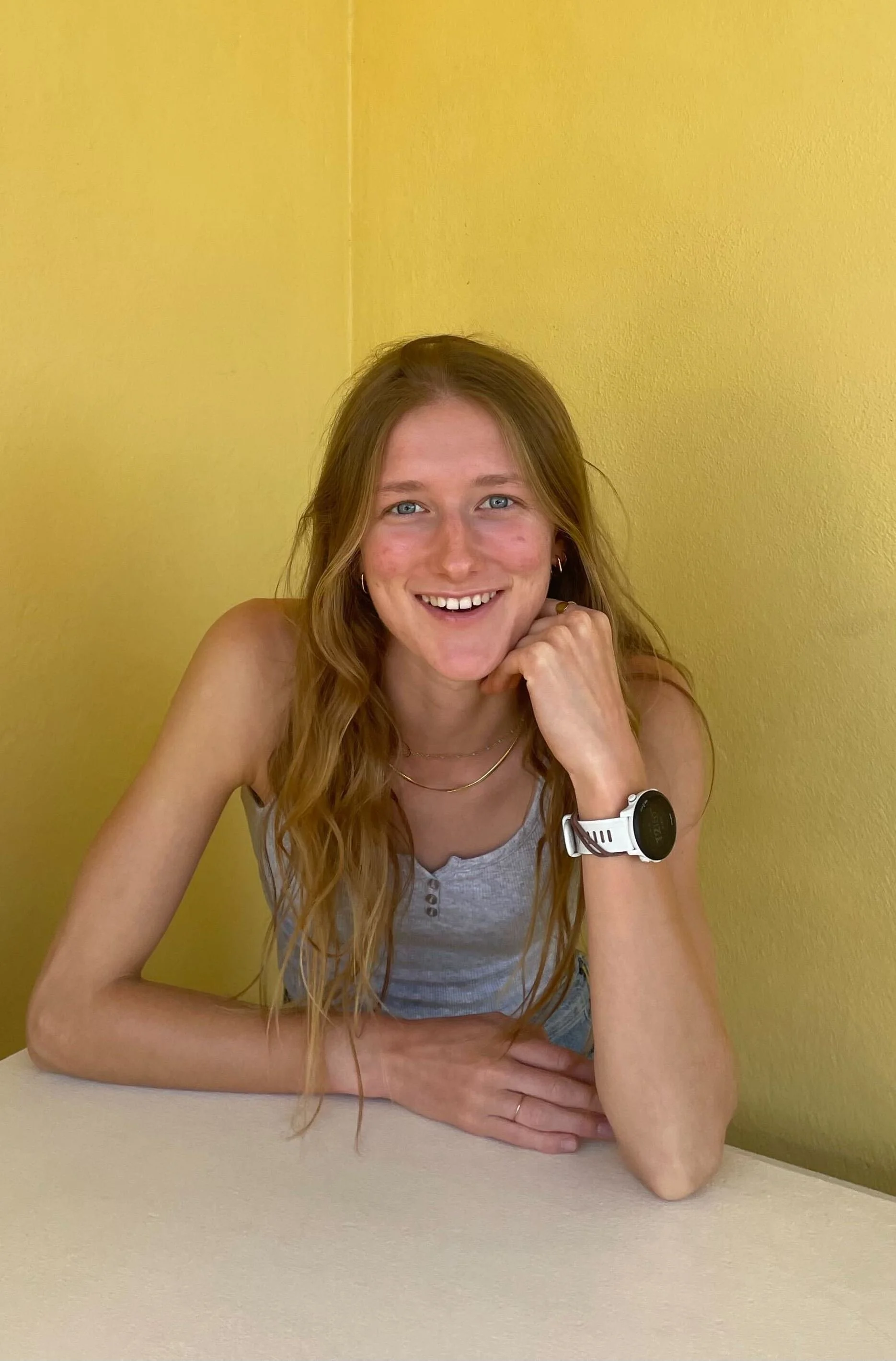 Smiling woman with long red hair, wearing a gray tank top, sitting at a table with a yellow wall behind her.